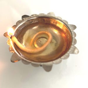 May include: A small, round, metallic oil lamp with a scalloped edge and a polished gold-toned interior. A single cotton wick is submerged in oil and lit, producing a small, bright flame that illuminates the center of the bowl-shaped vessel.
