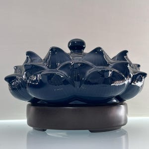 May include: A dark blue ceramic lotus flower-shaped essential oil diffuser with a glossy finish. The diffuser sits on a dark brown base and has a small, round knob on top. The design is reminiscent of a blooming lotus.