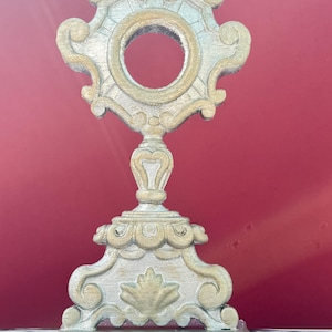 Puede incluir: Una custodia de color oro y plata que presenta un marco circular ornamentado con una abertura central, rematada con una cruz. La base es ancha y decorativa con motivos de hojas talladas, mostrando un acabado metálico con reflejos.