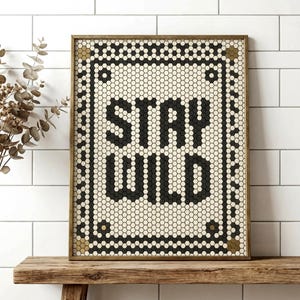 Könnte beinhalten: Gerahmter Kunstdruck mit dem Schriftzug "STAY WILD" in fetten schwarzen Buchstaben. Das Design zeigt ein mosaikartiges Muster aus schwarzen und cremefarbenen sechseckigen Fliesen, eingerahmt von einem goldfarbenen Rahmen. Das Kunstwerk wird auf einem rustikalen Holzregal präsentiert.