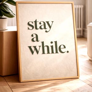 以下が含まれることがあります： 「stay a while」という言葉が濃い緑色の文字で書かれた額入りのプリント。アートワークは薄いベージュ色の背景で、明るい木材で額装されています。プリントは木の床の壁に立てかけられています。