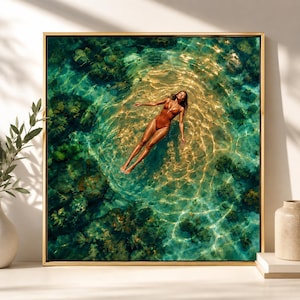 Puede incluir: Un cuadro cuadrado con marco dorado que muestra una vista aérea de una persona flotando en agua turquesa clara. La persona lleva un bañador de una pieza marrón, situada en una zona de luz solar dorada rodeada de rocas oscuras bajo el agua.