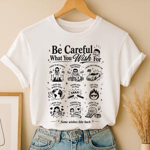 以下が含まれることがあります： 上部に「Be Careful What You Wish For」、下部に「Some wishes bite back」というテキストが黒いグラフィックでプリントされた白い半袖Tシャツ。デザインには、さまざまな願い事を描いた9つの小さなイラストが含まれています。