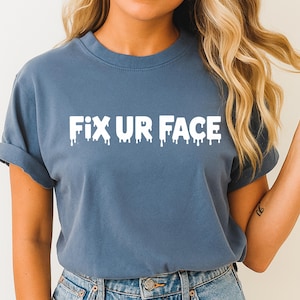 Könnte beinhalten: Ein staubblaues T-Shirt mit dem Text "FIX UR FACE" in weißer, tropfender Schrift. Das Shirt hat kurze Ärmel und einen Rundhalsausschnitt. Das Model trägt eine blaue Jeans.