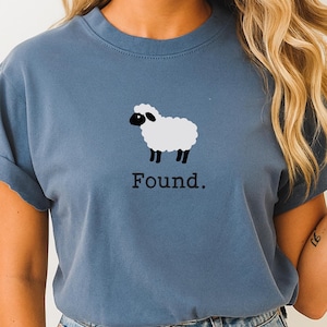 May include: A dusty blue crew neck t-shirt featuring a graphic of a white sheep with a black face and legs. Below the sheep illustration, the word "Found." is printed in a simple, dark, serif font. The shirt has short sleeves and a classic fit.