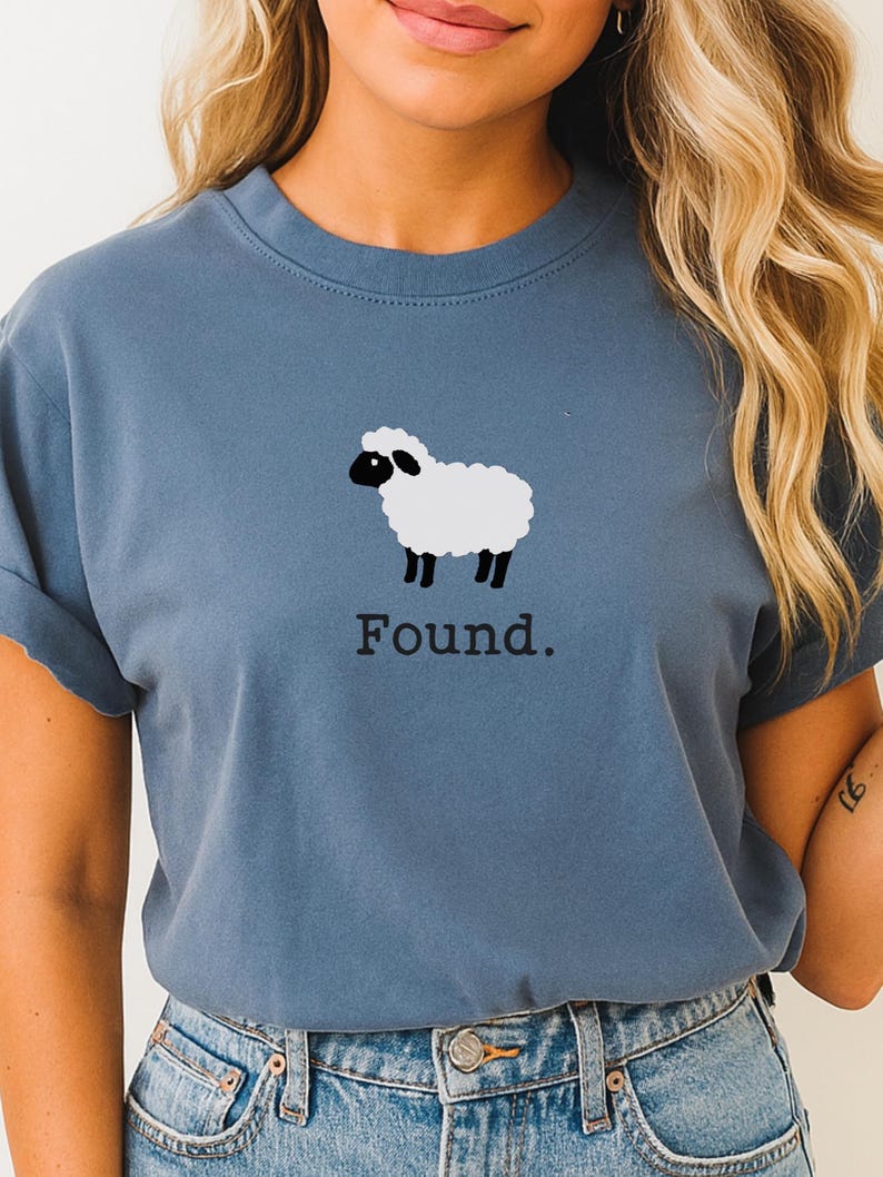 May include: A dusty blue crew neck t-shirt featuring a graphic of a white sheep with a black face and legs. Below the sheep illustration, the word "Found." is printed in a simple, dark, serif font. The shirt has short sleeves and a classic fit.