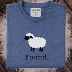 May include: A heather blue crew neck t-shirt featuring a graphic of a white sheep with a black face and legs. Below the sheep, the word "Found." is printed in a black serif font. The shirt has a Gildan Softstyle label indicating a size small.