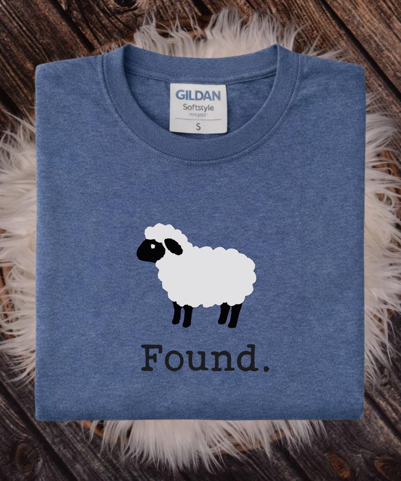 May include: A heather blue crew neck t-shirt featuring a graphic of a white sheep with a black face and legs. Below the sheep, the word "Found." is printed in a black serif font. The shirt has a Gildan Softstyle label indicating a size small.