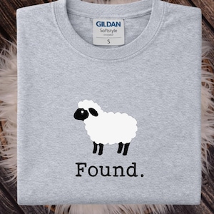 May include: A light gray crew neck t-shirt featuring a graphic of a white sheep with a black face and legs. Below the sheep, the word Found is printed in a black serif font. The shirt has a Gildan Softstyle label indicating a size small.