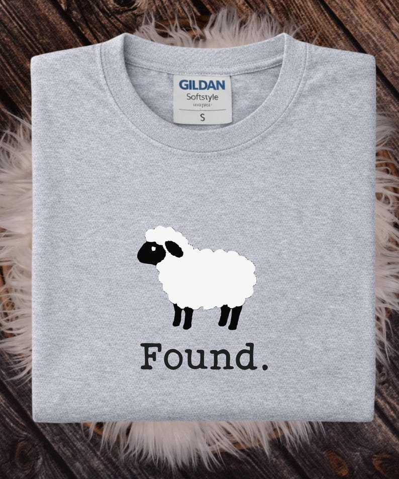 May include: A light gray crew neck t-shirt featuring a graphic of a white sheep with a black face and legs. Below the sheep, the word Found is printed in a black serif font. The shirt has a Gildan Softstyle label indicating a size small.