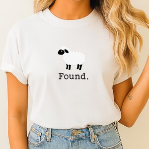 May include: A white short-sleeved t-shirt featuring a graphic of a white sheep with a black face and black legs. Below the sheep illustration, the word "Found." is printed in a black, serif-style font. The shirt has a crew neckline.