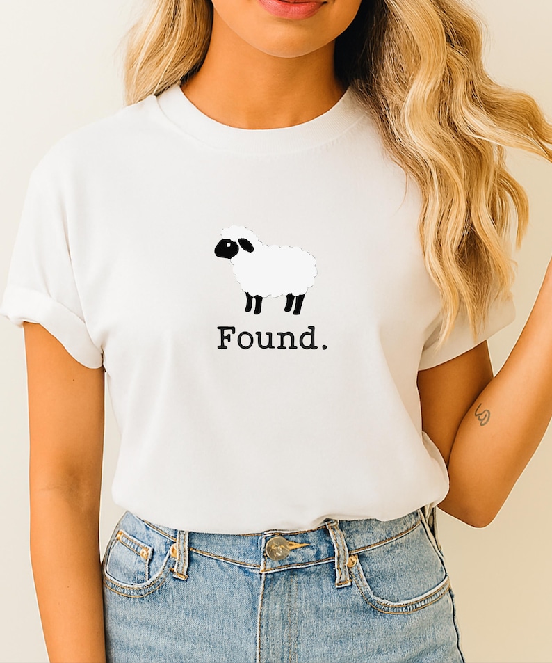 May include: A white short-sleeved t-shirt featuring a graphic of a white sheep with a black face and black legs. Below the sheep illustration, the word "Found." is printed in a black, serif-style font. The shirt has a crew neckline.