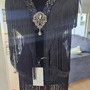 May include: Black fringed top with a decorative brooch. The top has long fringe, a sequined neckline, and a large, ornate brooch with a central stone and dangling pendant. The garment is displayed on a mannequin.