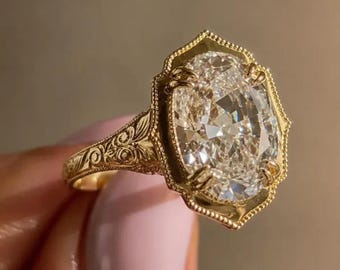 Puede incluir: Un anillo de oro con una gran piedra preciosa transparente de corte ovalado, engastada en un bisel decorativo de bordes festoneados. La banda de oro amarillo presenta intrincados patrones de volutas grabados en los laterales.