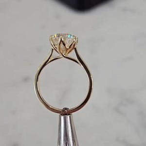 May include: A gold engagement ring featuring a large, round, clear gemstone. The ring has a distinctive setting with curved prongs that secure the stone. The band is polished, reflecting light. The ring is displayed against a blurred background.