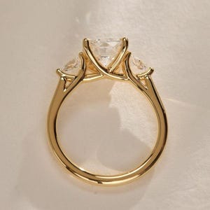 May include: A gold ring with a large, square-cut center stone and two smaller, triangular side stones. The ring has a polished gold band and a classic three-stone setting. The stones are clear and sparkling.