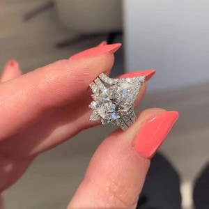 Könnte beinhalten: Ein funkelnder Diamantring mit einem großen, birnenförmigen Mittelstein und einem Halo aus kleineren Diamanten. Der Ring hat einen geteilten Schaft und zusätzliche Diamantakzente, die einen luxuriösen Look erzeugen.