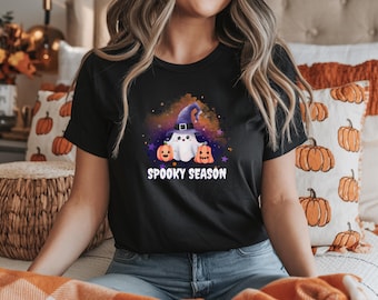 May include: Black t-shirt featuring a whimsical Halloween design. The graphic includes a ghost wearing a witch hat, two jack-o'-lanterns, and the words "Spooky Season" in white. The design is set against a colorful, starry background.