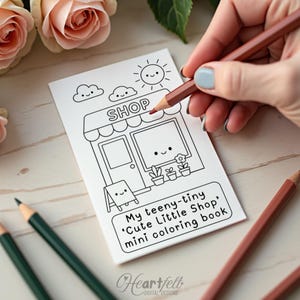 Pode incluir: Um mini livro de colorir branco com uma ilustração de uma loja, apresentando um sol sorridente, nuvens e uma fachada de loja. O texto diz "My teeny-tiny 'Cute Little Shop' mini coloring book." Uma mão está colorindo com um lápis marrom.