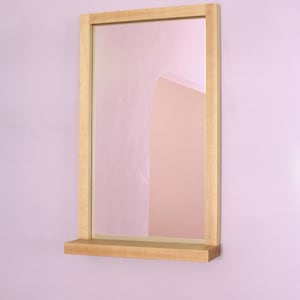 Pode incluir: Um espelho com moldura de madeira e uma prateleira embaixo. O espelho reflete uma parede rosa com uma porta arqueada.