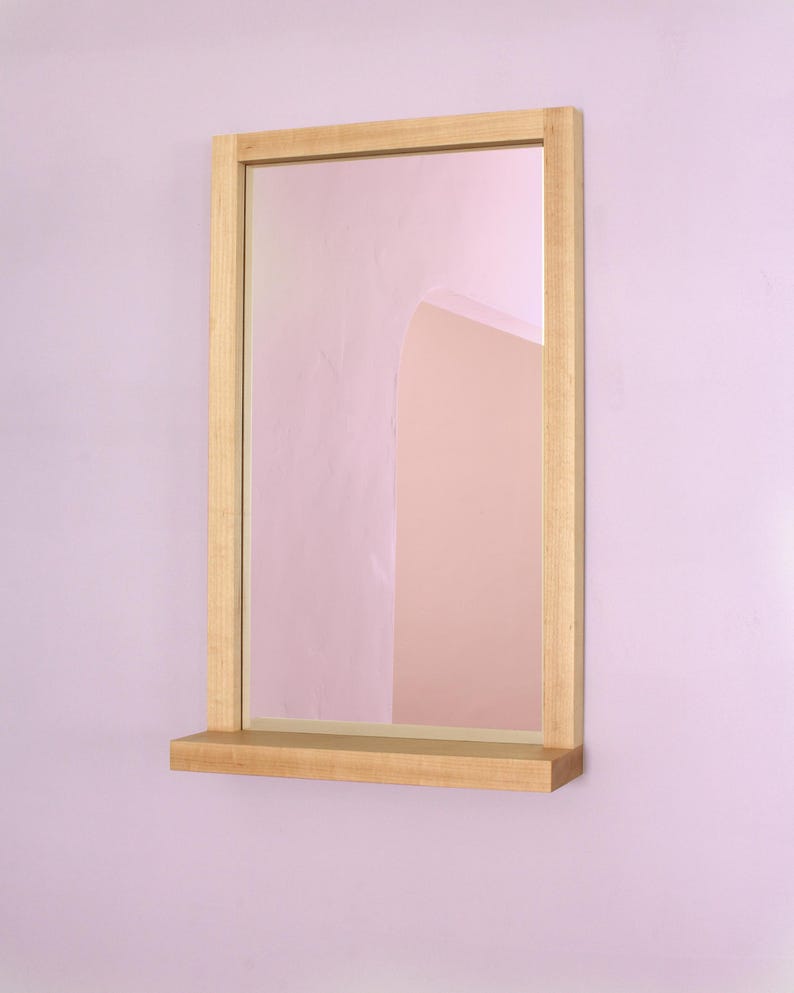 May include: A wooden framed mirror with a shelf underneath. The mirror is reflecting a pink wall with an arched doorway.