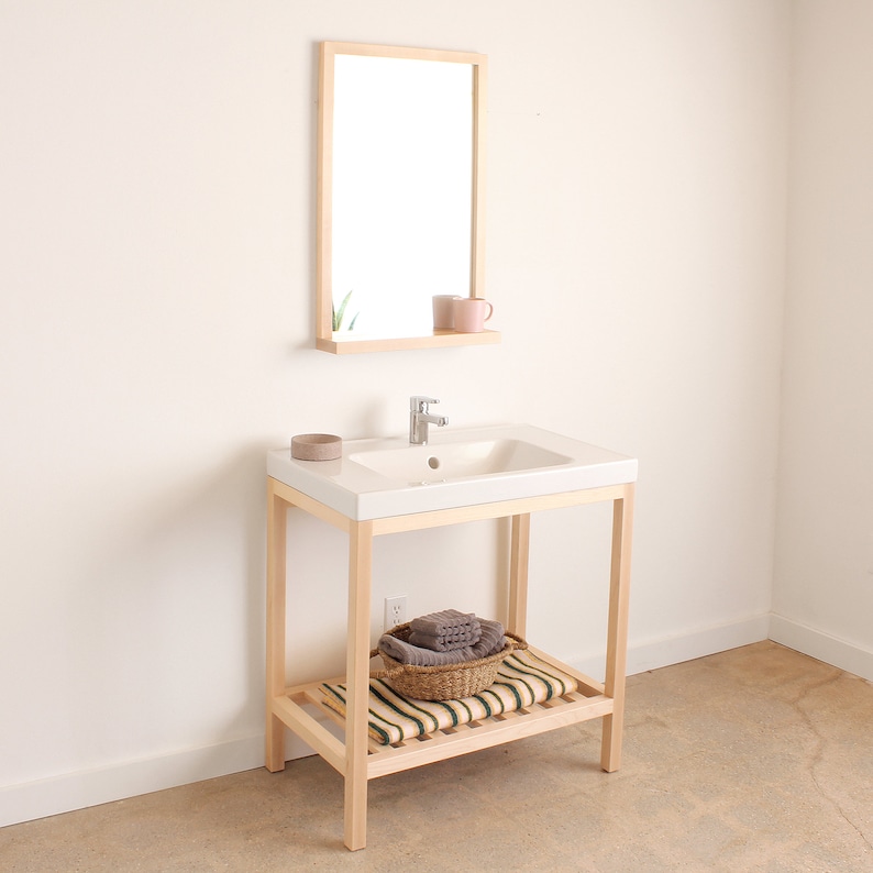 May include: A white bathroom vanity with a wooden frame and a white porcelain sink. The vanity has a shelf with a woven basket containing towels. A mirror with a wooden frame is mounted above the sink.