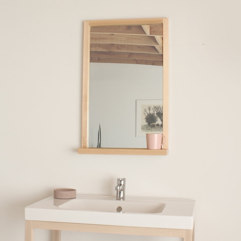 May include: A wooden framed mirror with a shelf underneath. The mirror is reflecting a room with a wooden ceiling and a picture on the wall. A pink mug is sitting on the shelf.