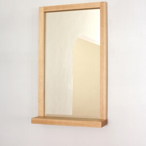 May include: A rectangular wooden mirror with a shelf underneath. The mirror has a light wood frame and the shelf is made of the same wood.