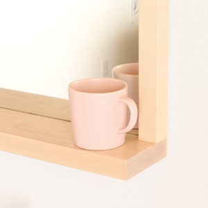May include: A light wood shelf with a pink mug on it. The shelf is attached to a mirror with a light wood frame.