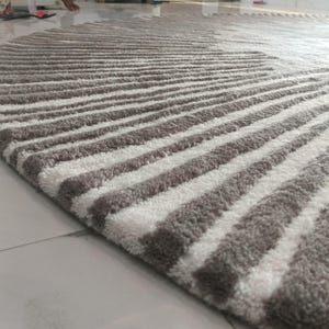 May include: A round area rug featuring a high-pile texture with alternating radial stripes in shades of taupe and white. The tufted pattern radiates from the center toward the curved edge, creating a consistent linear design across the surface.