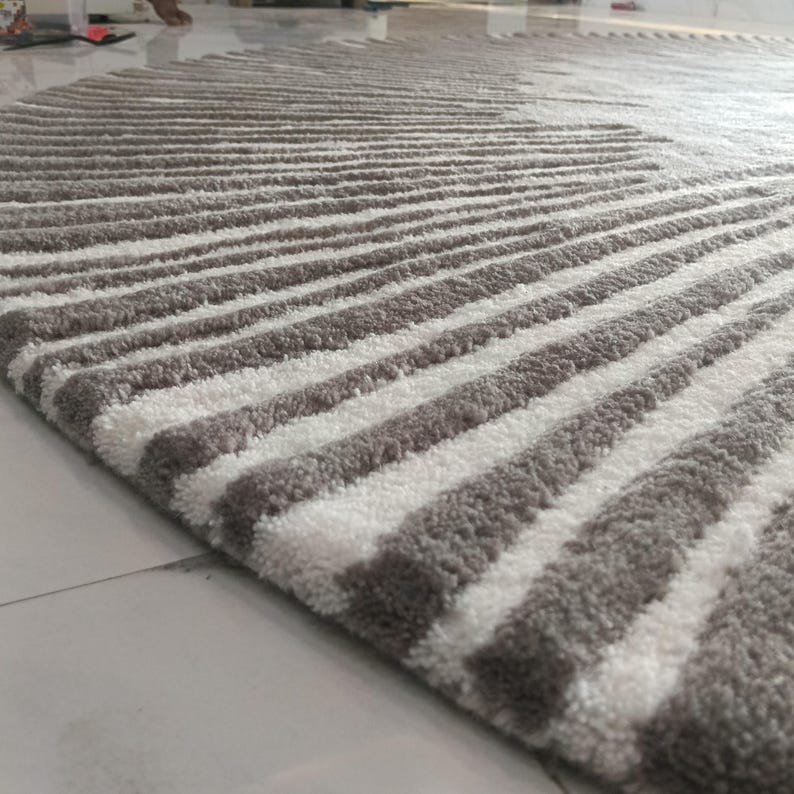 May include: A round area rug featuring a high-pile texture with alternating radial stripes in shades of taupe and white. The tufted pattern radiates from the center toward the curved edge, creating a consistent linear design across the surface.