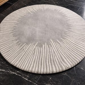 May include: A circular area rug featuring a solid light gray center surrounded by a wide border of radiating white lines. The design creates a sunburst pattern with varying lengths of white tufted lines extending outward from the central gray circle.