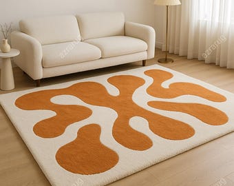 Op de afbeelding: Een rechthoekig, crèmekleurig vloerkleed met een abstract ontwerp in oranje tinten. Het vloerkleed heeft een zachte, pluche textuur en ligt op een houten vloer. Het ontwerp bestaat uit organische, amoebe-achtige vormen.