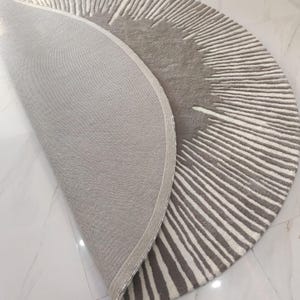 May include: A semi-circular rug featuring a textured pattern of radiating lines in shades of gray and white. The rug is partially folded over, revealing a solid gray fabric backing and a finished edge along the curved perimeter of the textile.