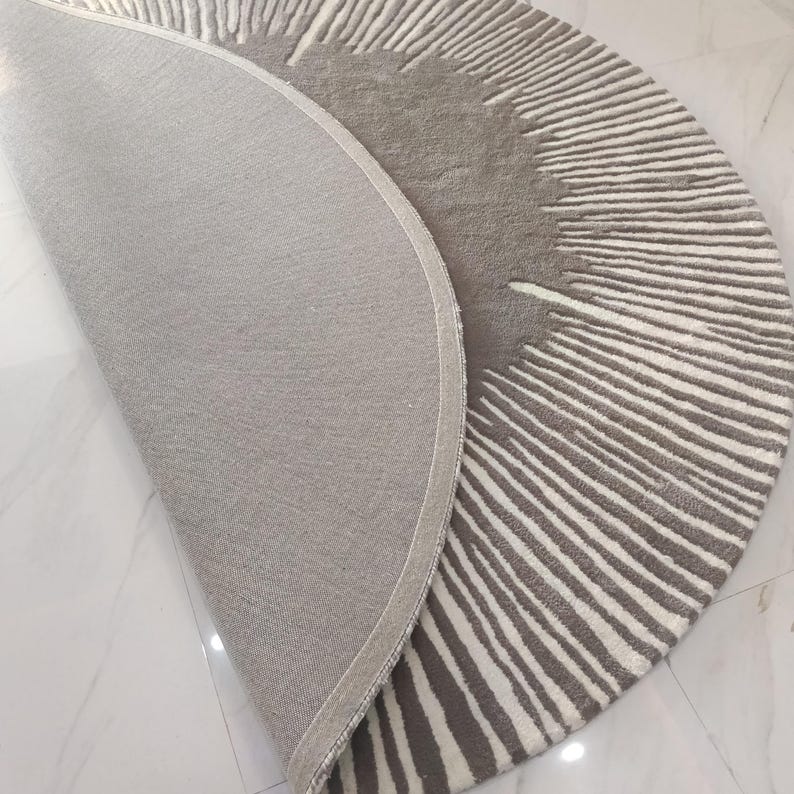 May include: A semi-circular rug featuring a textured pattern of radiating lines in shades of gray and white. The rug is partially folded over, revealing a solid gray fabric backing and a finished edge along the curved perimeter of the textile.