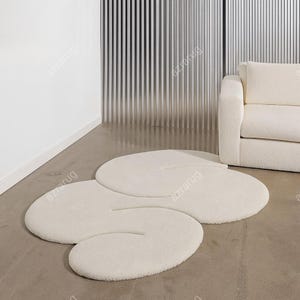 Op de afbeelding: Een crèmekleurig, abstract gevormd tapijt met een zacht, getextureerd oppervlak. Het ontwerp van het tapijt heeft drie met elkaar verbonden, ovale vormen. Het tapijt ligt op een betonnen vloer, met een witte bank en verticale panelen op de achtergrond.