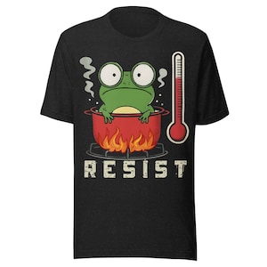 Peut inclure: T-shirt gris foncé avec une grenouille de dessin animé dans une marmite rouge sur des flammes, un thermomètre et le mot "RESIST". La grenouille a de grands yeux et de la vapeur qui monte. Un design humoristique.