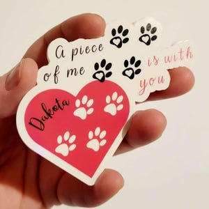 Puede incluir: Una pegatina en forma de coraz&oacute;n con el nombre "Dakota" en cursiva, rodeada de huellas de patas. La pegatina tambi&eacute;n presenta el texto "A piece of me is with you" en una fuente de escritura, con huellas de patas adicionales.