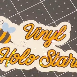 Puede incluir: Una pegatina blanca con las palabras "Vinyl Holo Stars" en escritura naranja. Una abeja de dibujos animados con rayas negras y amarillas y alas azules est&aacute; a la izquierda. La pegatina tiene un efecto brillante.