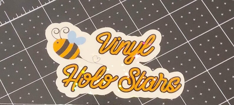 Puede incluir: Una pegatina blanca con las palabras "Vinyl Holo Stars" en escritura naranja. Una abeja de dibujos animados con rayas negras y amarillas y alas azules est&aacute; a la izquierda. La pegatina tiene un efecto brillante.