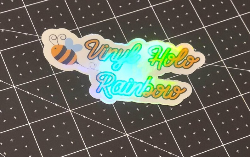 Puede incluir: Una pegatina hologr&aacute;fica de vinilo con las palabras "Vinyl Holo Rainbow" en una fuente de escritura. La pegatina presenta una abeja de dibujos animados con rayas naranjas y negras. La pegatina est&aacute; sobre una superficie negra con un patr&oacute;n de cuadr&iacute;cula.