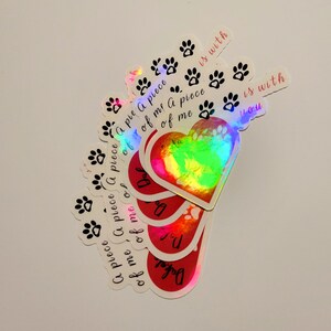 Puede incluir: Una colecci&oacute;n de pegatinas hologr&aacute;ficas con varios dise&ntilde;os. Algunas pegatinas presentan huellas de patas, mientras que otras tienen forma de coraz&oacute;n con el texto "A piece of me is with you". Las pegatinas tienen un acabado brillante e iridiscente.