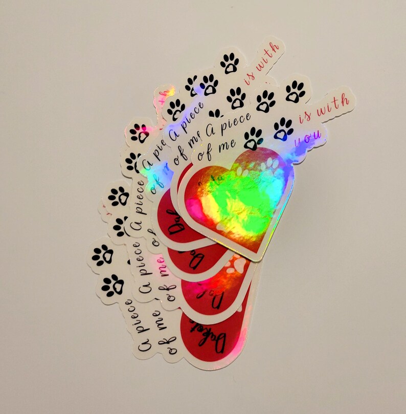 Puede incluir: Una colecci&oacute;n de pegatinas hologr&aacute;ficas con varios dise&ntilde;os. Algunas pegatinas presentan huellas de patas, mientras que otras tienen forma de coraz&oacute;n con el texto "A piece of me is with you". Las pegatinas tienen un acabado brillante e iridiscente.