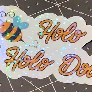 Puede incluir: Una pegatina hologr&aacute;fica con una abeja de dibujos animados y las palabras "Holo Holo Dots" en escritura naranja. La pegatina tiene un fondo blanco con efecto brillante, sobre una superficie negra.