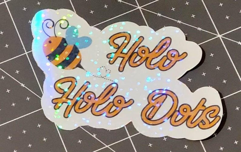 Puede incluir: Una pegatina hologr&aacute;fica con una abeja de dibujos animados y las palabras "Holo Holo Dots" en escritura naranja. La pegatina tiene un fondo blanco con efecto brillante, sobre una superficie negra.