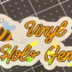 Puede incluir: Una pegatina hologr&aacute;fica con el texto "Vinyl Holo Gems" en fuente cursiva amarilla. La pegatina presenta una abeja de dibujos animados con rayas negras y amarillas y alas azules. Sobre una esterilla de corte negra.