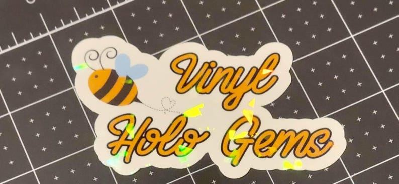 Puede incluir: Una pegatina hologr&aacute;fica con el texto "Vinyl Holo Gems" en fuente cursiva amarilla. La pegatina presenta una abeja de dibujos animados con rayas negras y amarillas y alas azules. Sobre una esterilla de corte negra.