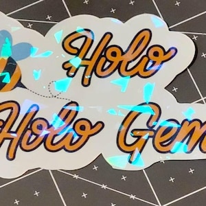 Puede incluir: Una pegatina hologr&aacute;fica con las palabras "Holo Holo Gems" en escritura naranja con un contorno negro. Una abeja de dibujos animados con rayas naranjas y negras y alas azules est&aacute; a la izquierda. La pegatina tiene un efecto brillante e iridiscente.