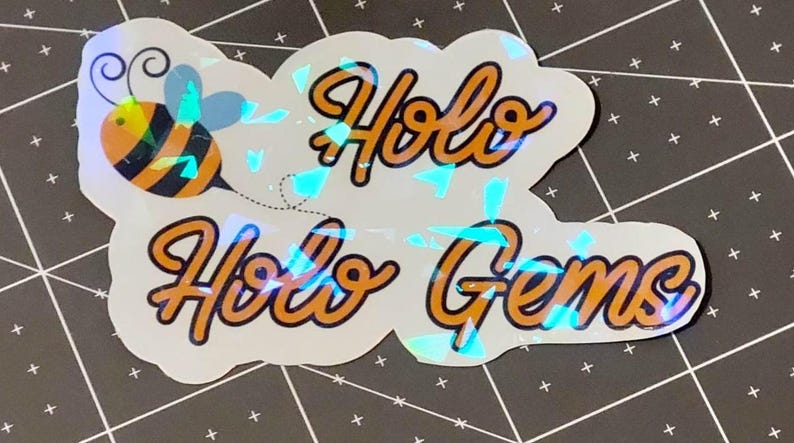 Puede incluir: Una pegatina hologr&aacute;fica con las palabras "Holo Holo Gems" en escritura naranja con un contorno negro. Una abeja de dibujos animados con rayas naranjas y negras y alas azules est&aacute; a la izquierda. La pegatina tiene un efecto brillante e iridiscente.