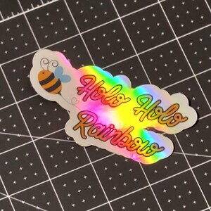 Puede incluir: Una pegatina hologr&aacute;fica con las palabras "Holo Holo Rainbows" en una fuente de escritura. La pegatina tiene un degradado de arco&iacute;ris y una ilustraci&oacute;n de abeja de dibujos animados. La pegatina est&aacute; sobre una esterilla de corte negra.